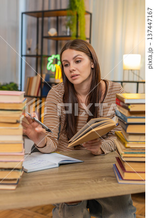 Young woman with stack of books writes notes from book looks very drained yet keeps focus on study Young woman with stack of books writes notes from book looks very drained yet keeps focus on study 137447167
