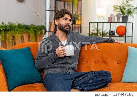 Indian man with hot tea or coffee warms hands gazes out window thoughtful look soft smile calm mood Indian man with hot tea or coffee warms hands gazes out window thoughtful look soft smile calm mood 137447169