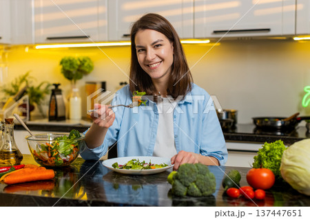 Young woman eating healthy salad in kitchen smiling with fork during nutrition lifestyle meal break 137447651
