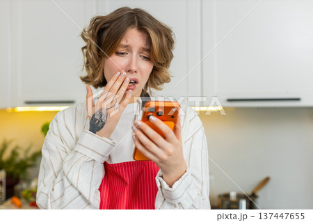 Young woman checking smartphone with worried reaction in kitchen during healthy meal making at home 137447655