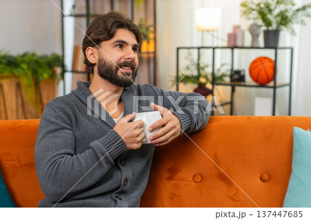 Indian man with hot tea or coffee warms hands gazes out window thoughtful look soft smile calm mood Indian man with hot tea or coffee warms hands gazes out window thoughtful look soft smile calm mood 137447685