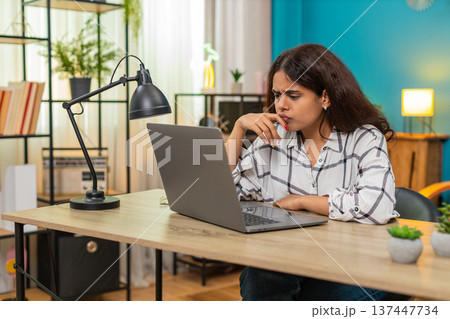 Indian woman saddened by emotional scene on laptop covering lips in shock sorrow indoors table desk 137447734