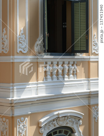Sharp shadows play across the warm, light-colored facade, emphasizing the Neoclassical depth of the scrollwork. Amidst the architectural texture, an open window with dark green shutters stands as a Sharp shadows play across the warm, light-colored facade, emphasizing the Neoclassical depth of the scrollwork. Amidst the architectural texture, an open window with dark green shutters stands as a 137451940