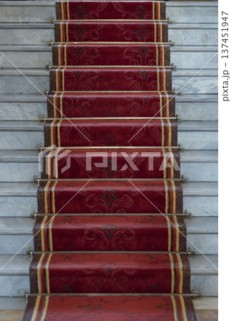 This high-angle shot captures a stone staircase featuring white marble treads and risers. Centered on the flight is a plush crimson runner accented with ornate gold motifs and decorative borders. Each This high-angle shot captures a stone staircase featuring white marble treads and risers. Centered on the flight is a plush crimson runner accented with ornate gold motifs and decorative borders. Each 137451947