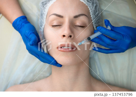 Female head in medical cap lying on a couch and get professional cosmetic injection in nasolabial fold area, aesthetic medicine procedure with doctor hands in blue gloves, close-up Female head in medical cap lying on a couch and get professional cosmetic injection in nasolabial fold area, aesthetic medicine procedure with doctor hands in blue gloves, close-up 137453796
