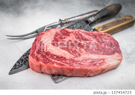 Premium raw ribeye beef steak with intense marbling and thick fat cap. Rustic butcher style on light stone background. 137454723