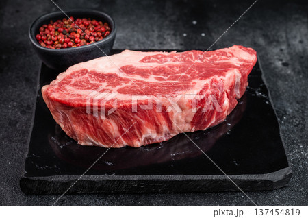 Raw beef chuck eye steak with rich marbling on stone board with pepper and fork. Fresh beef cut overhead on dark background. Raw beef chuck eye steak with rich marbling on stone board with pepper and fork. Fresh beef cut overhead on dark background. 137454819