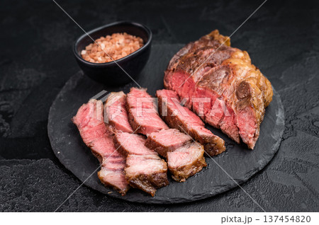Sliced beef chuck eye roll steak cooked medium rare with juicy texture. Gourmet beef meat with salt on dark stone background. Sliced beef chuck eye roll steak cooked medium rare with juicy texture. Gourmet beef meat with salt on dark stone background. 137454820