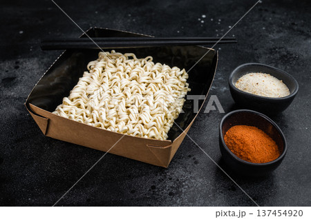 Dry instant ramen noodle block in black bowl with chopsticks and seasoning on dark background top view Asian convenience food concept Dry instant ramen noodle block in black bowl with chopsticks and seasoning on dark background top view Asian convenience food concept 137454920