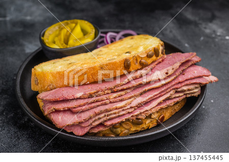 Smoked pastrami brisket slices in a hearty sandwich with seed crusted bread, onions and pickles on dark stone surface. 137455445