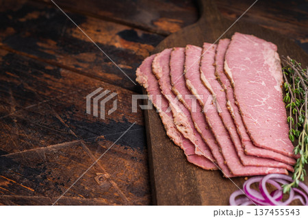 Smoked pastrami slices on a wooden board with a forged knife, red onion rings and fresh thyme. Premium deli beef presented on a dark rustic background, ideal for cooking or serving. 137455543