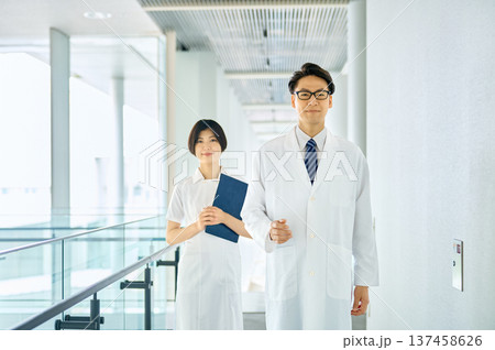 病院の廊下を歩く笑顔の日本人男性医師と女性看護師。チーム医療と信頼のイメージ。 病院の廊下を歩く笑顔の日本人男性医師と女性看護師。チーム医療と信頼のイメージ。 137458626