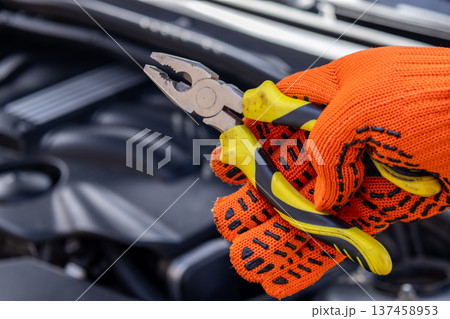 A mechanic is focused on fixing a car engine while holding pliers in one hand. The setting is a garage filled with tools A mechanic is focused on fixing a car engine while holding pliers in one hand. The setting is a garage filled with tools 137458953