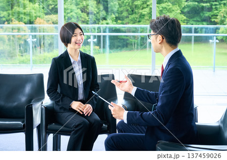 明るいオフィスのロビーで採用面接を受けるリクルートスーツの女性と面接官のビジネスマ 明るいオフィスのロビーで採用面接を受けるリクルートスーツの女性と面接官のビジネスマ 137459026