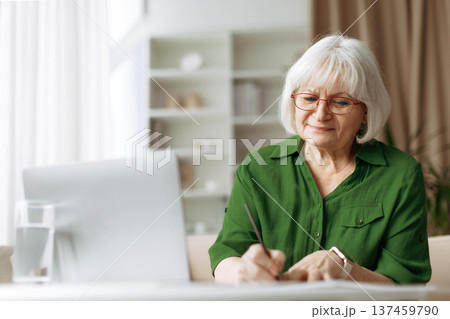 Senior woman writing notes while using laptop at home Senior woman writing notes while using laptop at home 137459790