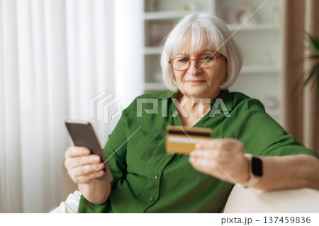 Senior woman sitting on sofa using smartphone and holding credit card Senior woman sitting on sofa using smartphone and holding credit card 137459836