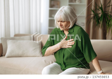 Senior woman touching chest while sitting on sofa at home Senior woman touching chest while sitting on sofa at home 137459853