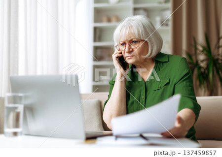 Serious senior woman reviewing document at home while speaking on phone 137459857