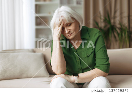 Senior woman sitting on sofa with worried expression and holding head 137459873