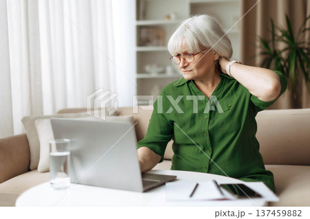 Senior woman touching neck with discomfort while sitting at laptop at home Senior woman touching neck with discomfort while sitting at laptop at home 137459882