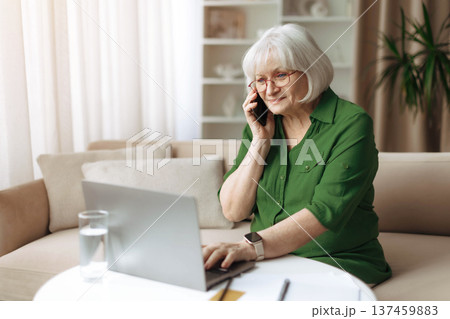 Senior woman talking on smartphone while working on laptop at home 137459883