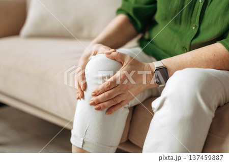 Close up of senior woman holding painful knee while sitting on sofa at home Close up of senior woman holding painful knee while sitting on sofa at home 137459887