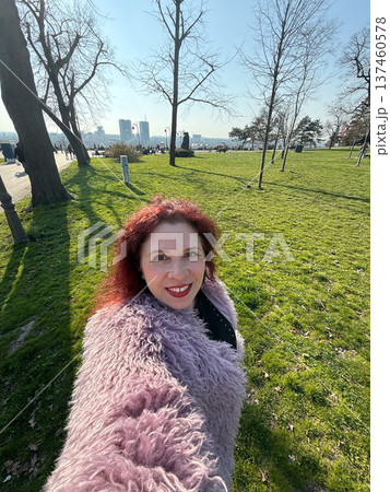 Smiling woman taking selfie in sunny park with green grass and trees. Outdoor lifestyle, spring atmosphere and joyful urban recreation moment. Smiling woman taking selfie in sunny park with green grass and trees. Outdoor lifestyle, spring atmosphere and joyful urban recreation moment. 137460578