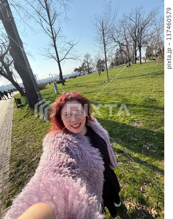 Smiling woman taking selfie in sunny city park with green lawn and trees. Outdoor lifestyle moment, spring atmosphere and joyful urban recreation. 137460579
