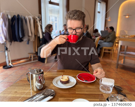 Man drinking coffee or tea from red cup while sitting at wooden table in cafe interior. Relaxed urban lifestyle, coffee culture and casual social dining moment. 137460600