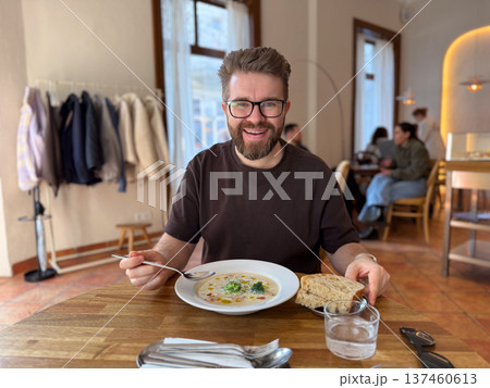 Smiling man sitting at wooden table with soup and bread in bright cafe. Friendly dining moment, urban cafe culture, everyday lifestyle and relaxed social atmosphere. 137460613
