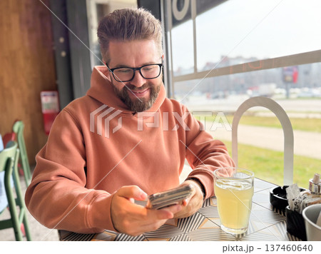 Smiling man using smartphone while sitting in a cafe near a large window with a drink on the table. Leisure lifestyle, digital communication, remote work culture, relaxation and modern urban social Smiling man using smartphone while sitting in a cafe near a large window with a drink on the table. Leisure lifestyle, digital communication, remote work culture, relaxation and modern urban social 137460640