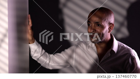 Confident businessman leaning against frosted glass in dark room with striped shadows 137460770