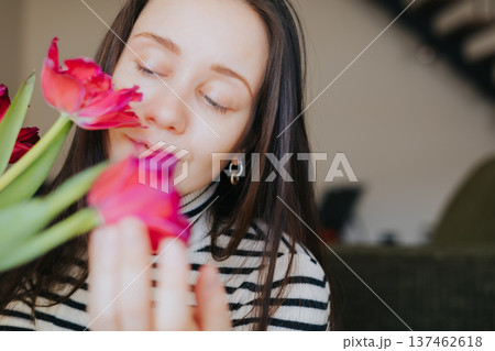 Young woman admiring red tulips bouquet at home. Gentle aesthetic of spring morning and self care. Blurred flowers in foreground. Concept of mindfulness, seasonal joy, and natural female beauty. Young woman admiring red tulips bouquet at home. Gentle aesthetic of spring morning and self care. Blurred flowers in foreground. Concept of mindfulness, seasonal joy, and natural female beauty. 137462618