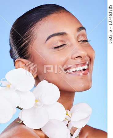 Skincare, flower and happy woman in studio for beauty, wellness and organic body care on blue background. Floral, face and Indian female model smile with orchid for eco friendly dermatology or shine Skincare, flower and happy woman in studio for beauty, wellness and organic body care on blue background. Floral, face and Indian female model smile with orchid for eco friendly dermatology or shine 137463285