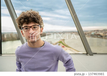 Young man with curly hair and glasses wearing a purple sweater, looking directly at the camera with a blurred panoramic city and river view behind him Young man with curly hair and glasses wearing a purple sweater, looking directly at the camera with a blurred panoramic city and river view behind him 137463981
