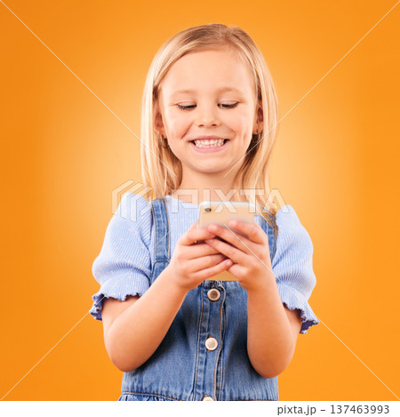 Child, smile and typing with phone in studio for social media, play mobile games and download app on orange background. Happy girl kid, smartphone and reading notification, watch multimedia and chat Child, smile and typing with phone in studio for social media, play mobile games and download app on orange background. Happy girl kid, smartphone and reading notification, watch multimedia and chat 137463993