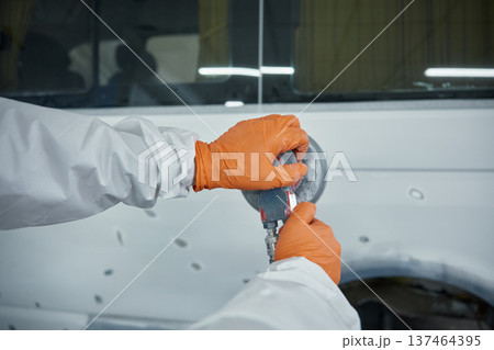 An auto body technician sands a car panel in a protective suit and orange gloves An auto body technician sands a car panel in a protective suit and orange gloves 137464395