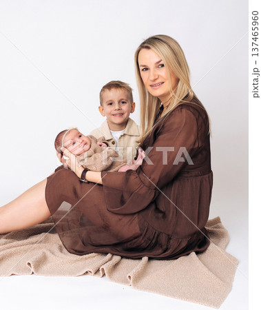 Young mother sitting on the floor holding her newborn baby while older son stands nearby. Warm family scene showing motherhood, parenting and love between family members.  137465960