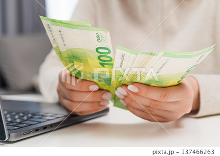 Woman counting euro banknotes while typing notes on laptop. Personal finance, budgeting, online accounting, small business management and money planning concept. 137466263