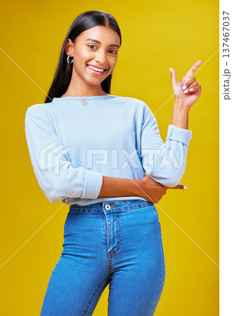Pointing, mockup and portrait of a woman in a studio for advertising, marketing or promotion. Smile, happy and young Indian female model with a direction hand gesture isolated by yellow background. Pointing, mockup and portrait of a woman in a studio for advertising, marketing or promotion. Smile, happy and young Indian female model with a direction hand gesture isolated by yellow background. 137467037