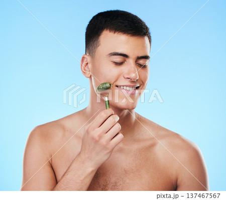 Man, beauty and face with skincare, roller for skin and grooming with smile on blue background. Jade crystal, model and dermatology, cosmetic tools and treatment with facial massage in a studio Man, beauty and face with skincare, roller for skin and grooming with smile on blue background. Jade crystal, model and dermatology, cosmetic tools and treatment with facial massage in a studio 137467567