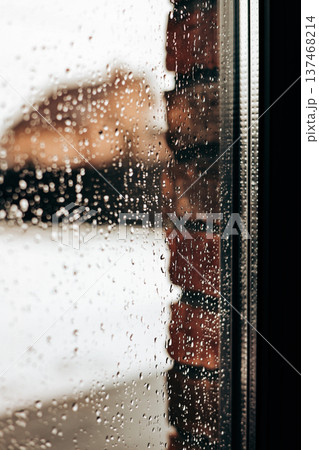 Raindrops on a window pane with a blurred brick wall background. Moody autumn or spring weather atmosphere. Texture of water drops on glass. Concept of loneliness, city rain, and peaceful solitude. 137468214