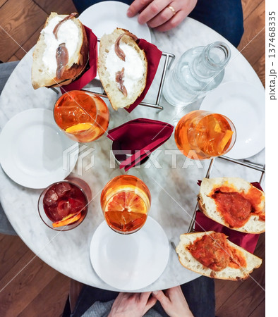 Overhead view of an italian aperitivo table featuring refreshing spritz cocktails, a dark red drink, water, and various appetizers including anchovy and meatball sandwiches Overhead view of an italian aperitivo table featuring refreshing spritz cocktails, a dark red drink, water, and various appetizers including anchovy and meatball sandwiches 137468335