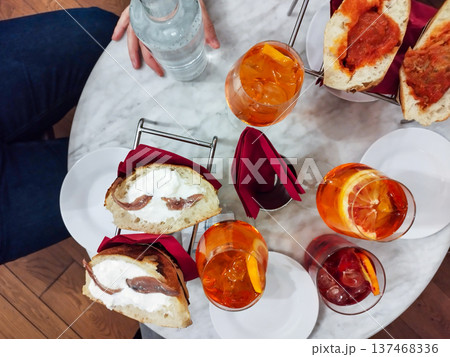 People enjoying a traditional italian aperitivo on a marble table, featuring refreshing orange spritz cocktails, red drinks, and various open-faced sandwiches with savory toppings in turin, italy 137468336