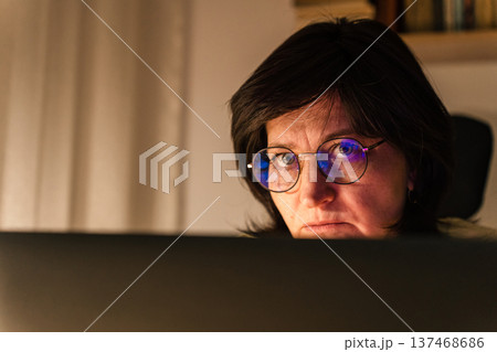 Woman wearing glasses with screen reflections on her eyes, focusing intently while working on laptop in dark environment night Woman wearing glasses with screen reflections on her eyes, focusing intently while working on laptop in dark environment night 137468686