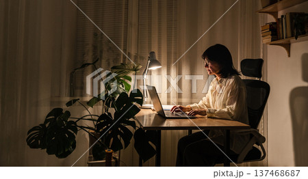 Woman focusing on work while sitting desk with laptop lamp illuminating her home office workspace, working on laptop late night Woman focusing on work while sitting desk with laptop lamp illuminating her home office workspace, working on laptop late night 137468687