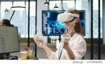 Female technician wearing VR goggles coding with AI language model assistance in startup office. Young woman using virtual reality integrating LLM assistant suggestions into code editor, camera A Female technician wearing VR goggles coding with AI language model assistance in startup office. Young woman using virtual reality integrating LLM assistant suggestions into code editor, camera A 137469262
