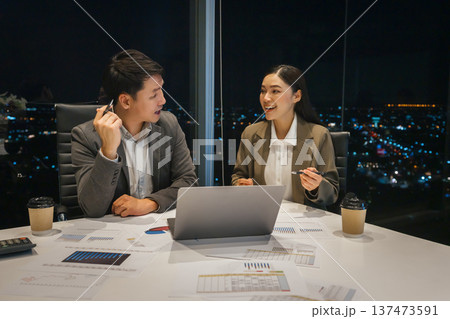 Two business colleagues talking while working with laptop computer together late in office with city lights background at night 137473591