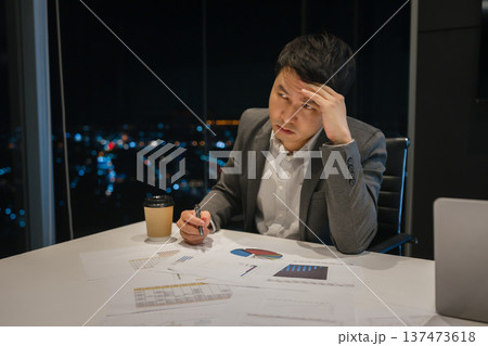 tired businessman looking worried and stressed while reviewing paperwork alone at night in office with city lights background tired businessman looking worried and stressed while reviewing paperwork alone at night in office with city lights background 137473618