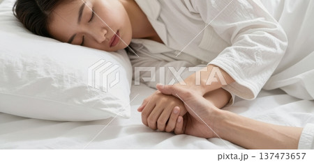 Close-up of a young Asian couple affectionately holding hands while the woman sleeps peacefully in a comfortable bed with white sheets. 137473657